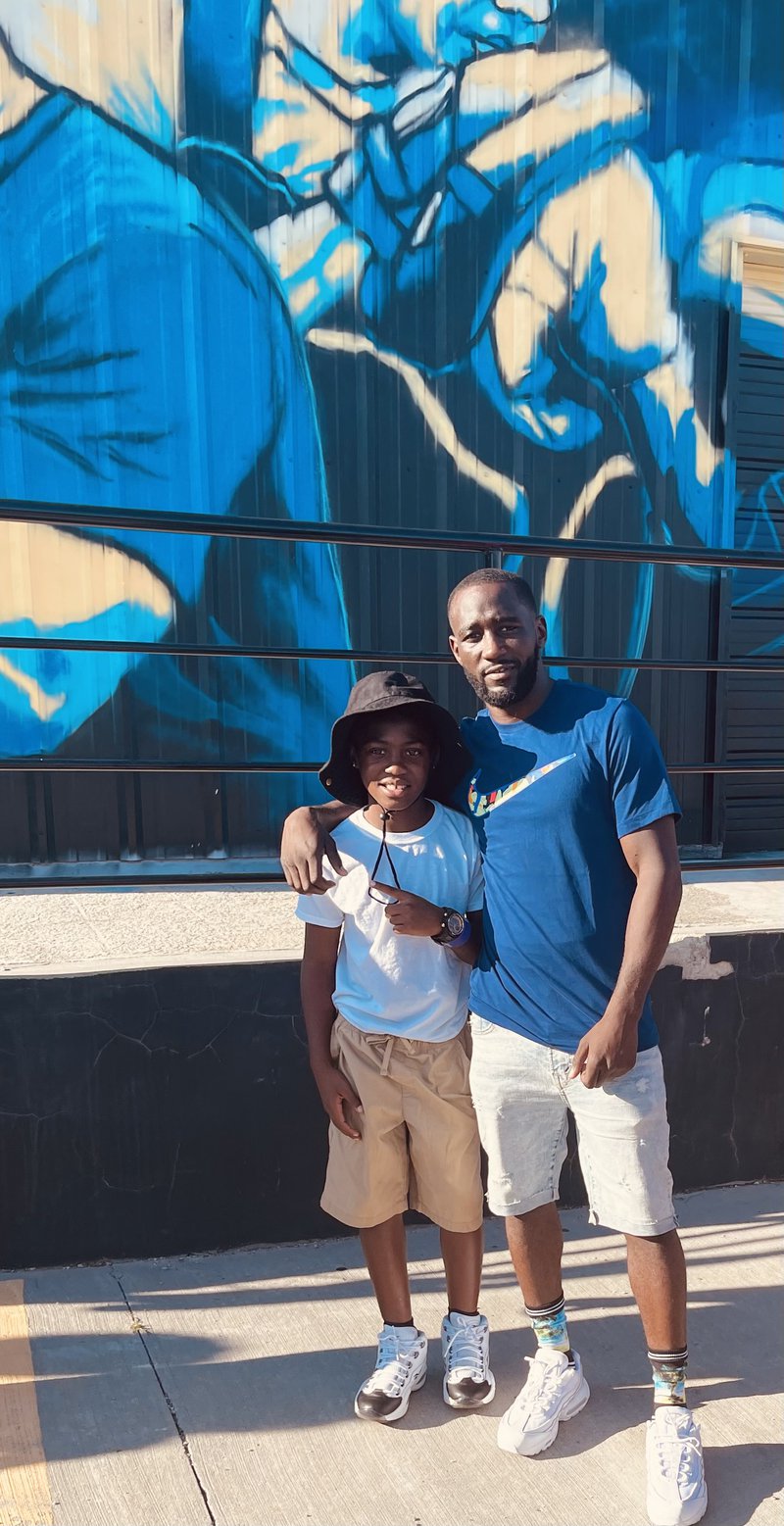 Bud Crawford with his arm around young Reno Busby standing in front a brightly-colored mural on the outside of a building.